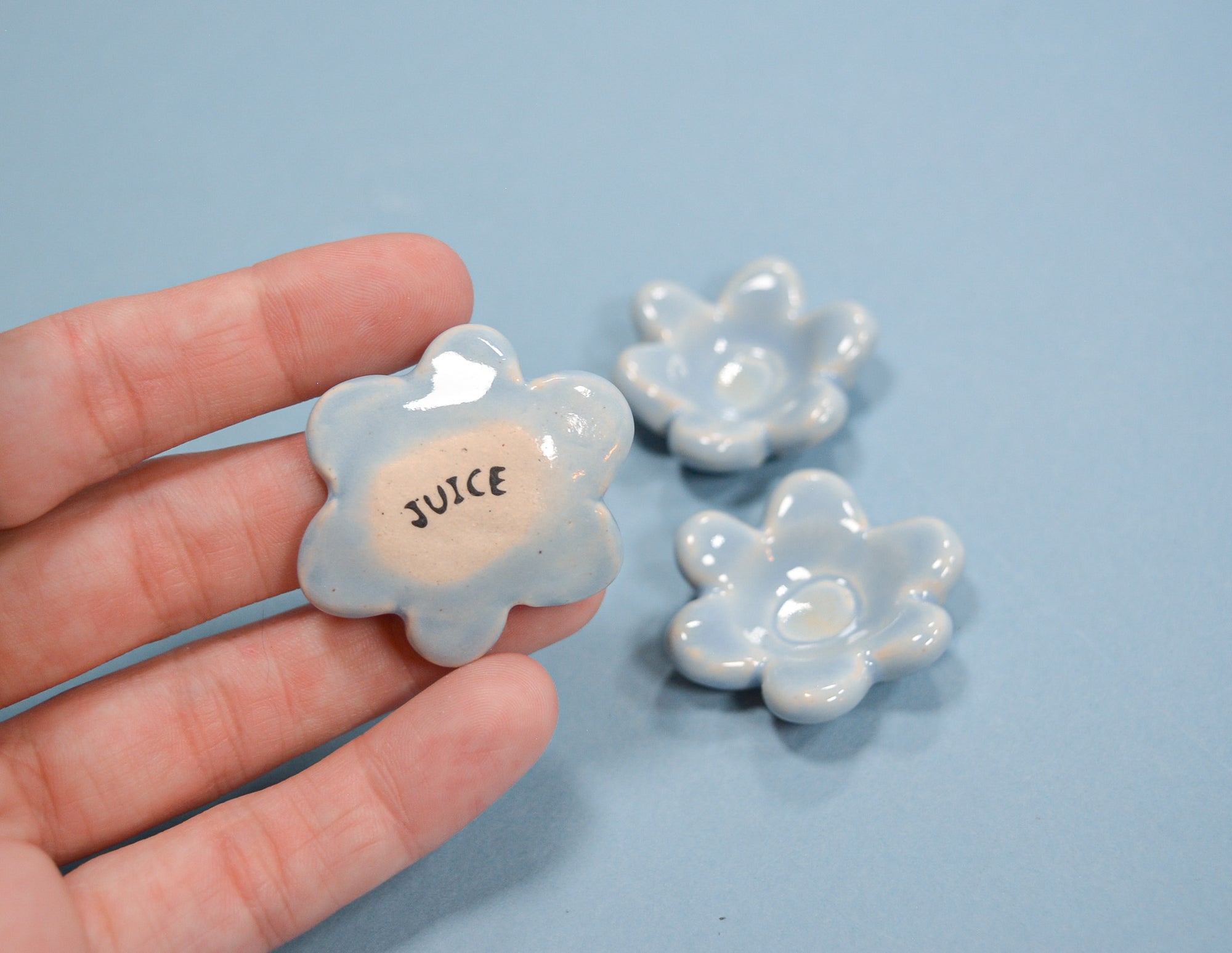 Three small flower-shaped ceramic condiment dishes in a glossy pale blue glaze, with one dish being held by a hand to show the bottom labeled 'JUICE'.
