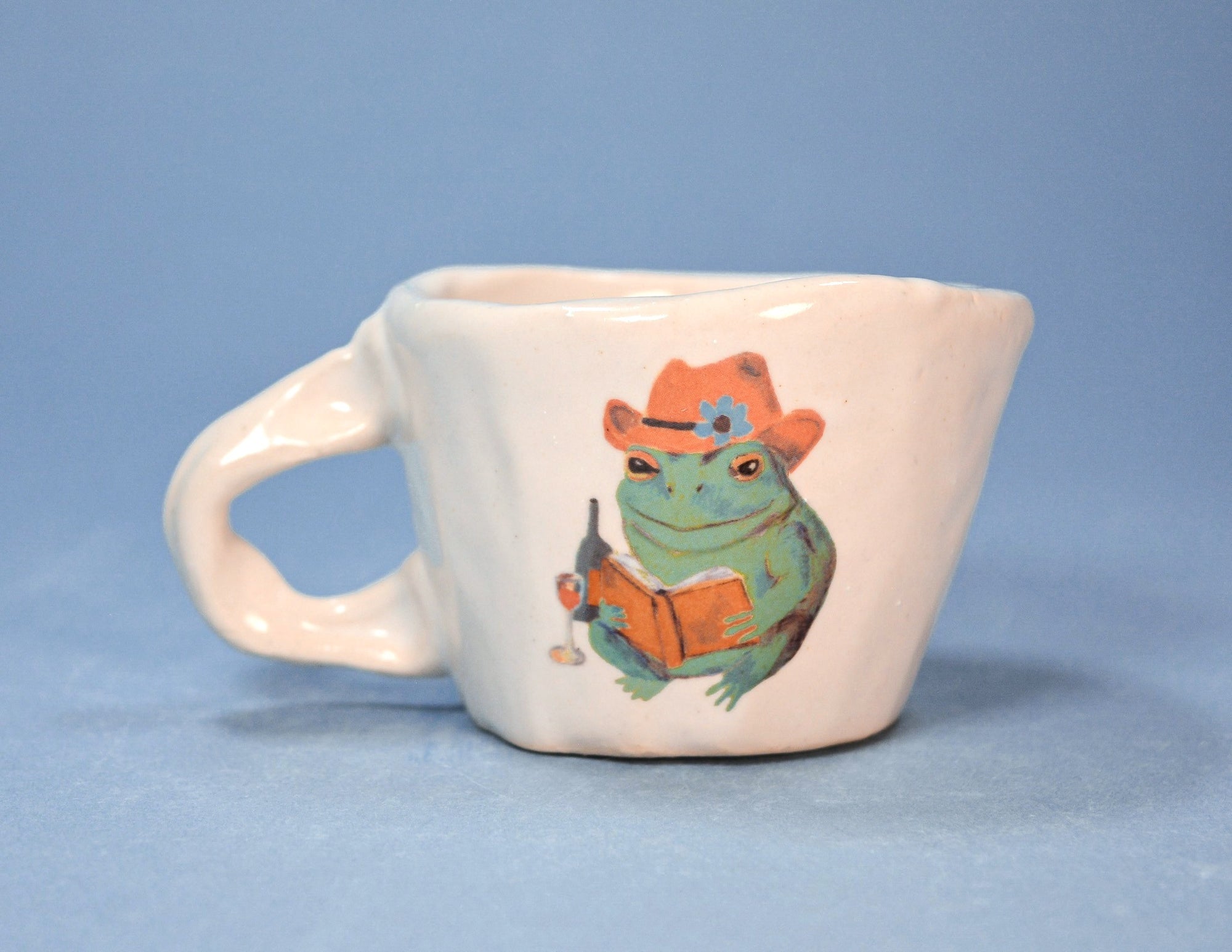 White mug with a frog wearing a cowboy hat and reading a book design on a blue background