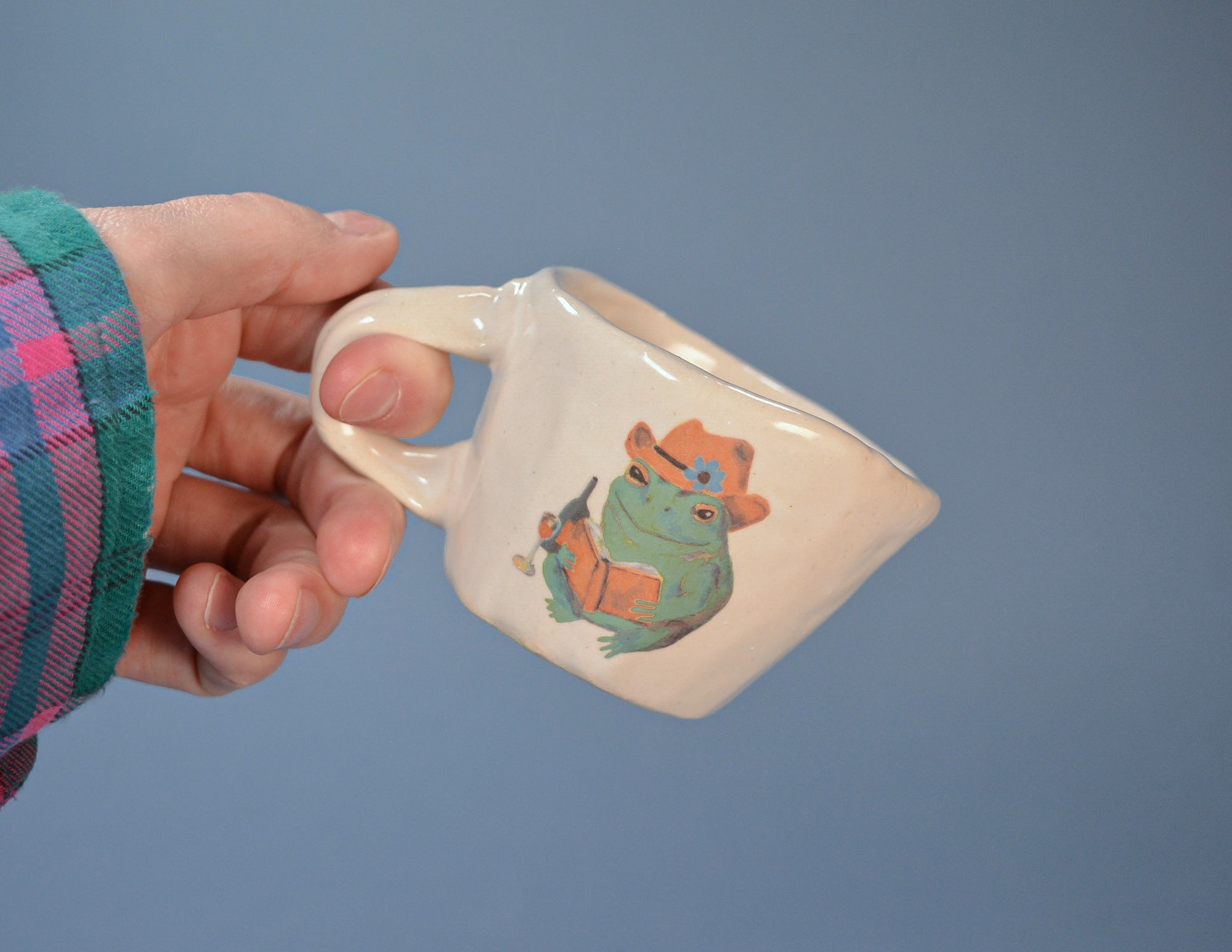 Hand holding a ceramic mug with a frog design against a blue background