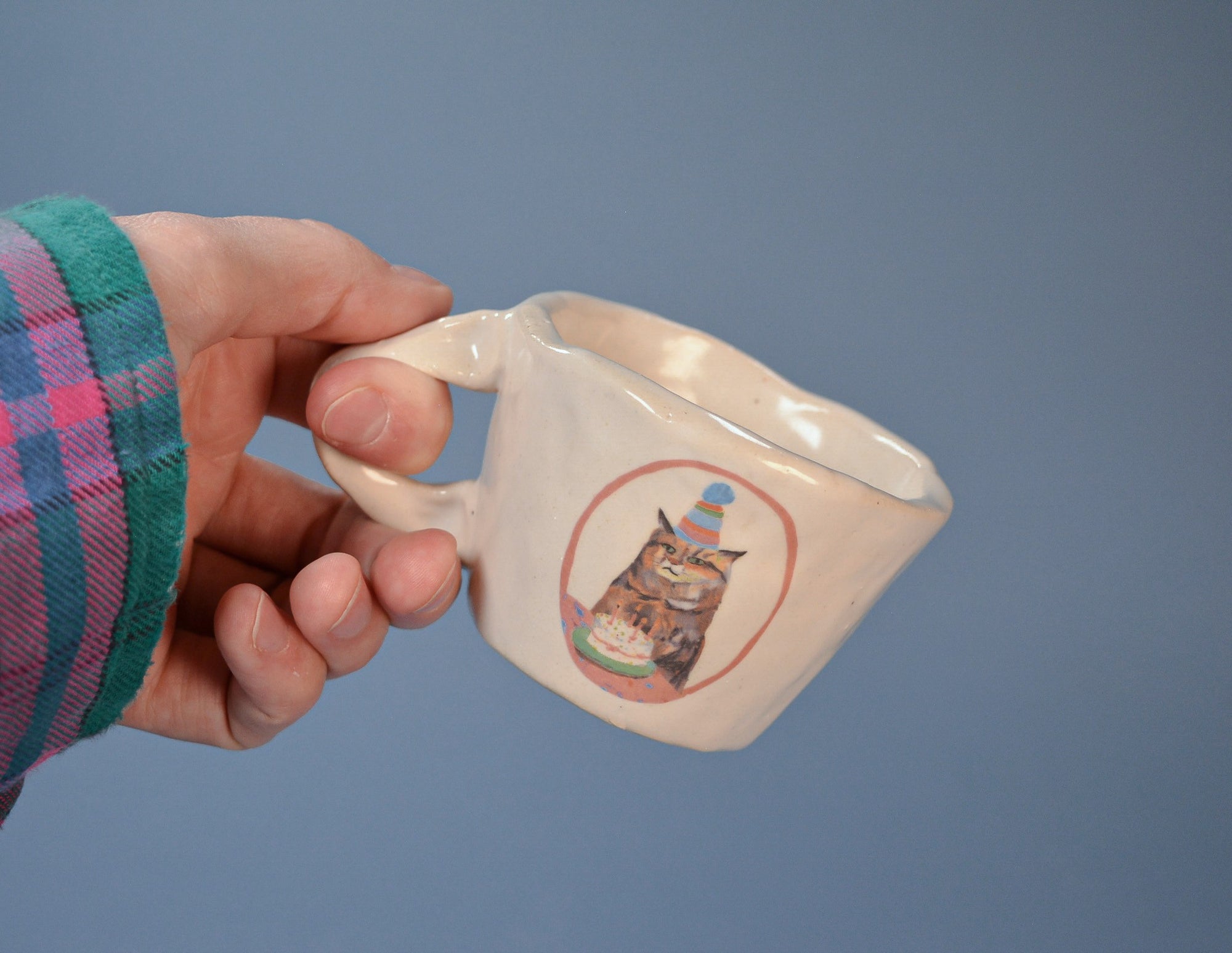 Hand holding a white ceramic mug with a cat illustration on a blue background
