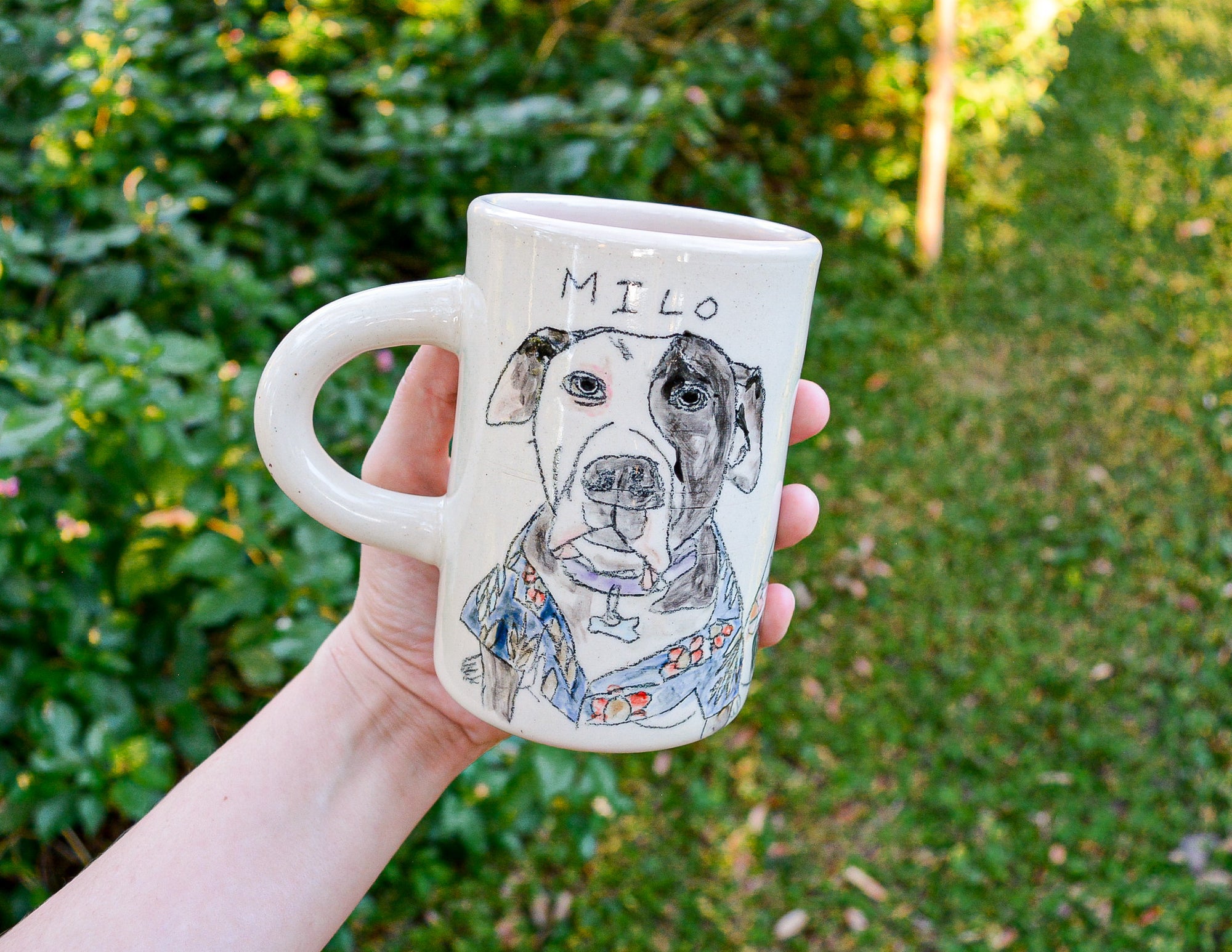 Person holding a mug with a dog illustration and 'Milo' text, outdoors.