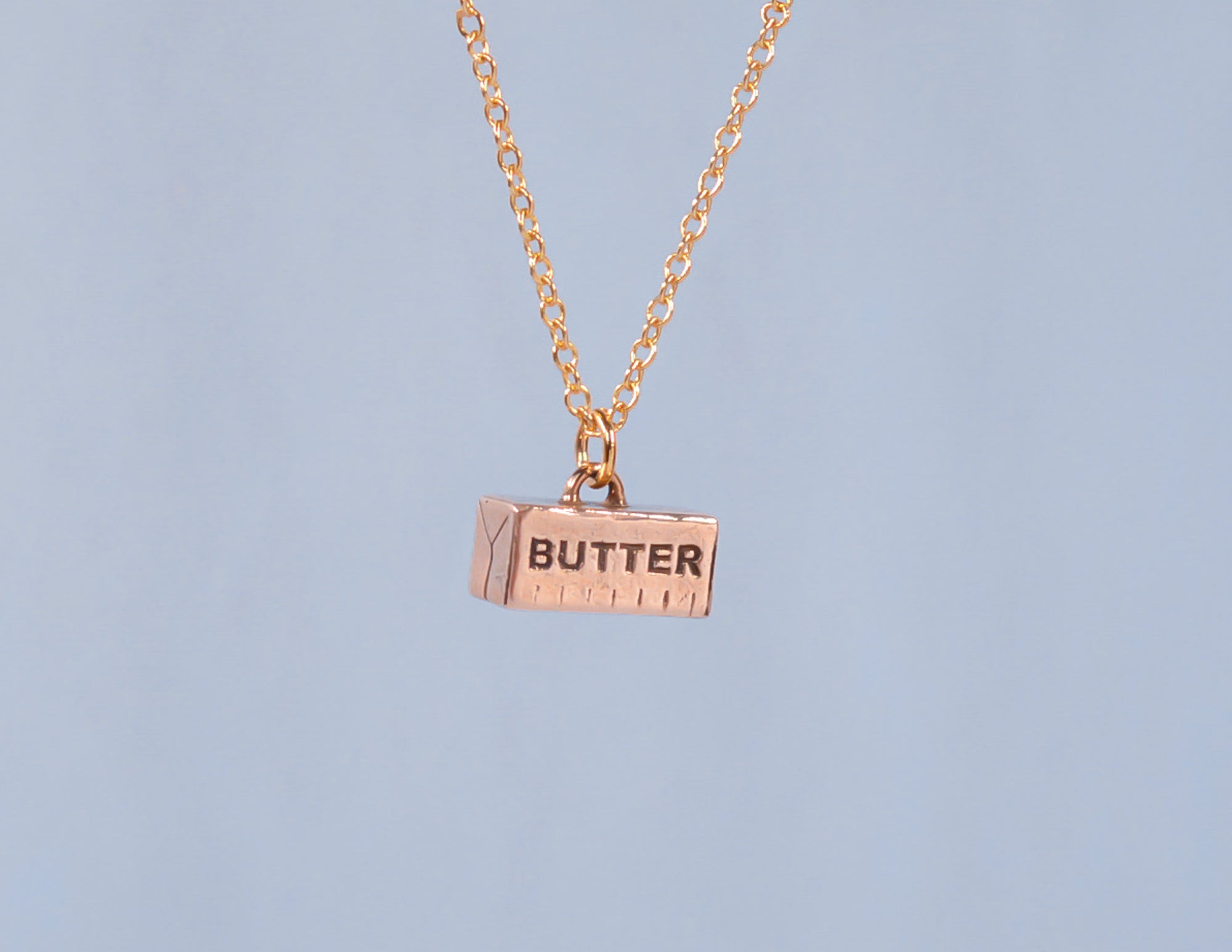 A bronze necklace in the shape of a stick of butter, with the word 'BUTTER' engraved on it, suspended on a 14k gold fill chain against a blue background.