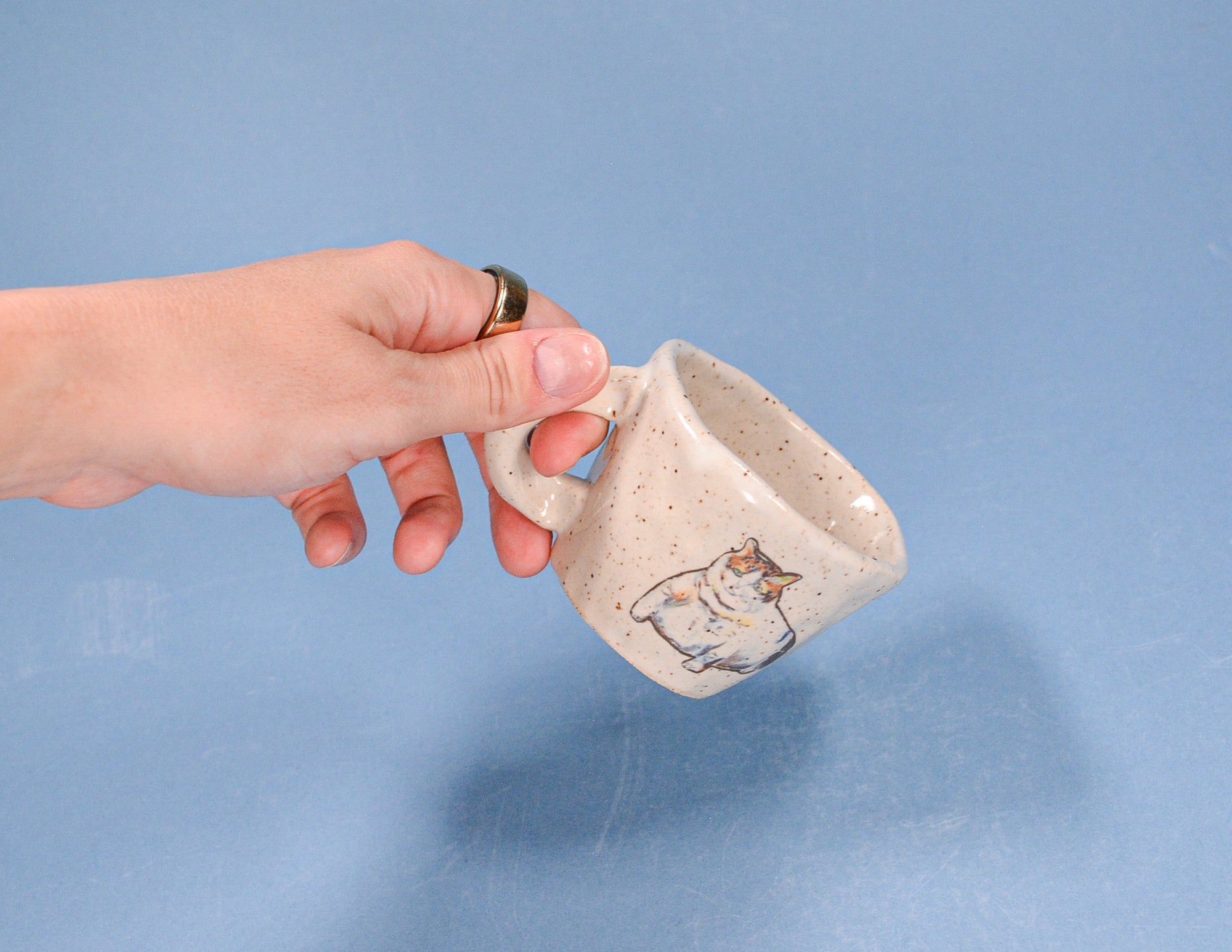 Hand holding a small ceramic mug with a cat design on a blue background