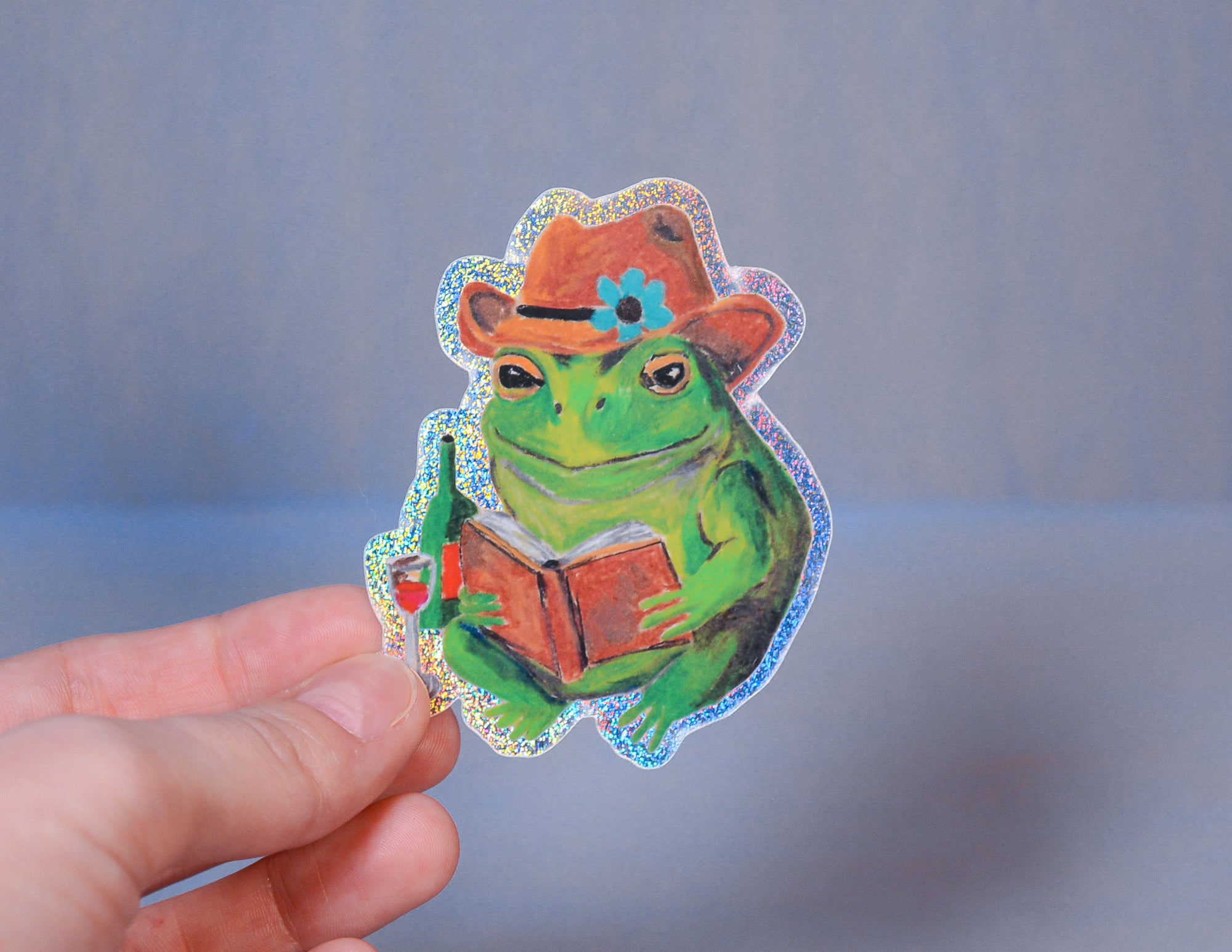 Sticker of a frog wearing a hat holding a book, held by a hand against a gray background