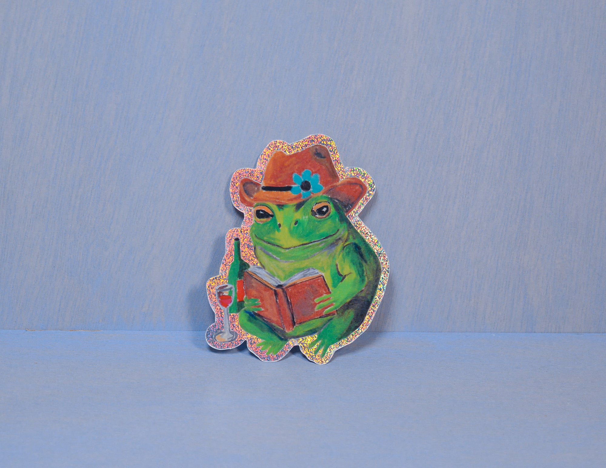Frog sticker wearing a cowboy hat and reading a book on a blue background