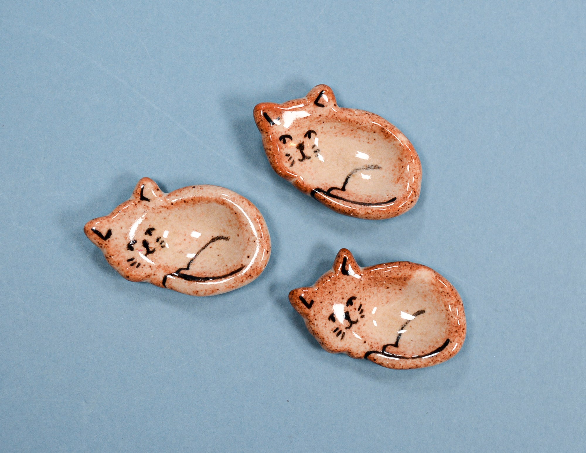 Three small ceramic cat-shaped dish holders with a speckled beige and terracotta glaze, decorated with black line drawings of sleeping cats, arranged on a solid blue background.