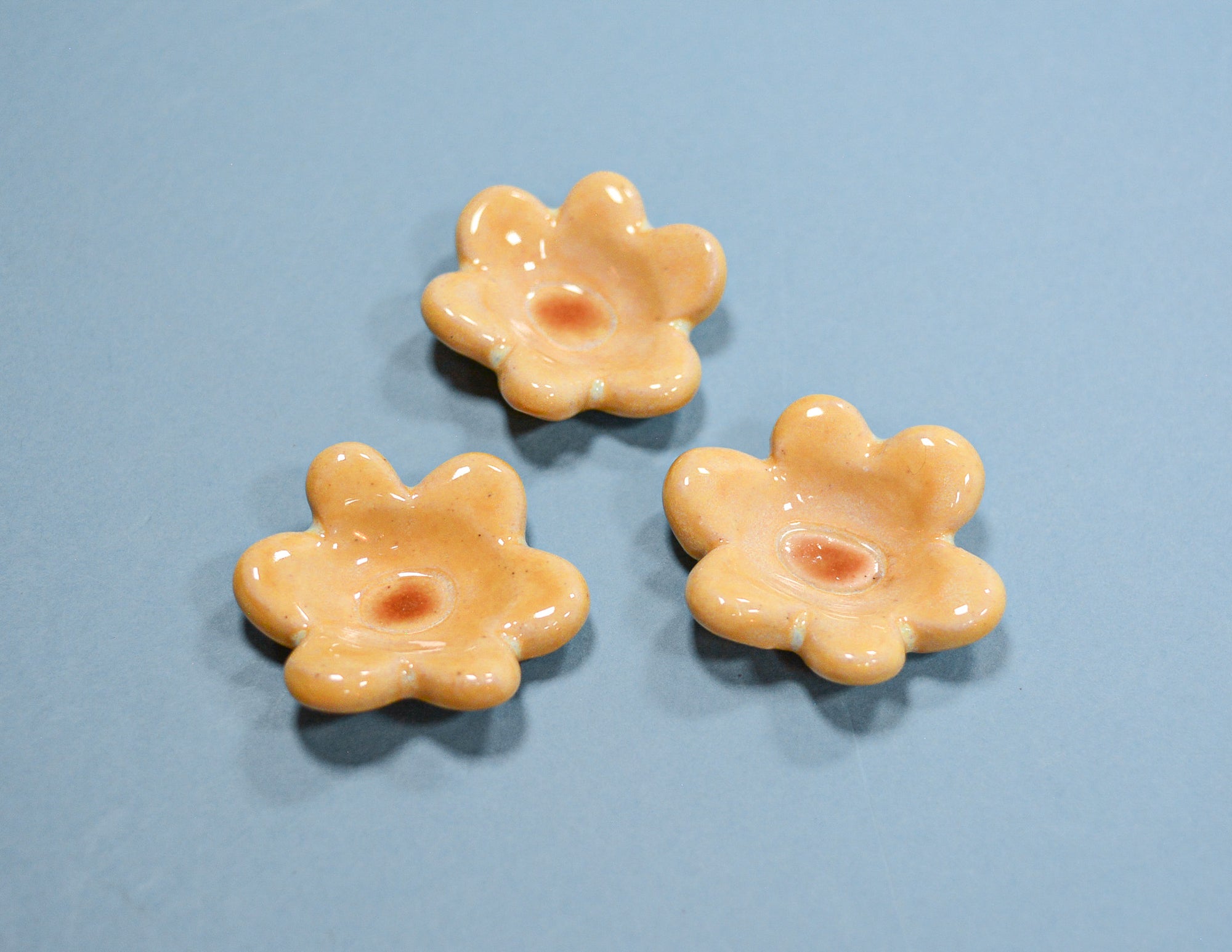 Three small glossy flower-shaped beads in a pale peachy orange color, arranged on a solid light blue background.